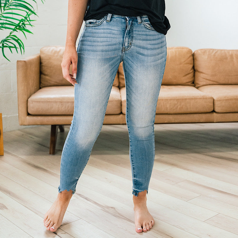 A person wearing light blue cropped jeans standing barefoot in front of a tan sofa on a light wood floor.