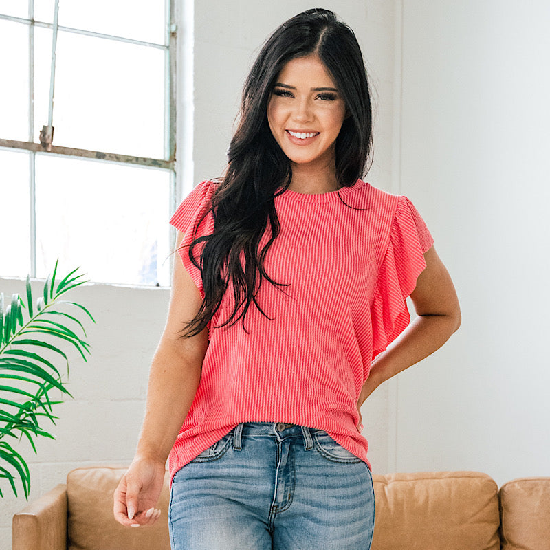 A woman smiling in a coral blouse and blue jeans while standing near a window, tan sofa, and green plant.