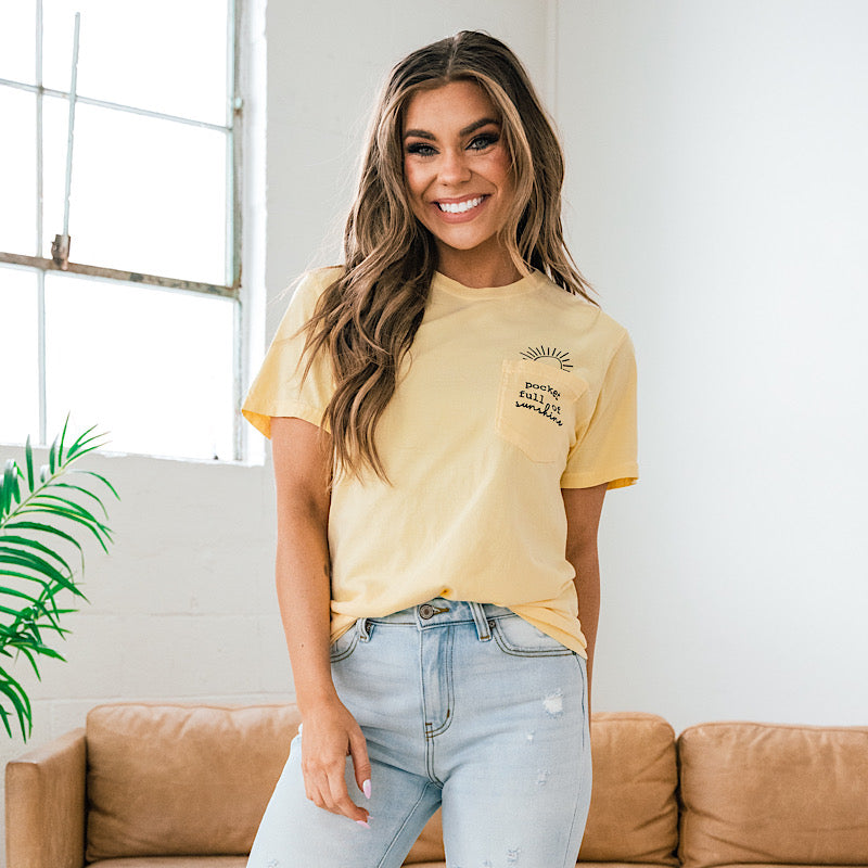 A woman wearing a yellow T-shirt and light jeans standing near a tan couch, white wall, window, and green plant.